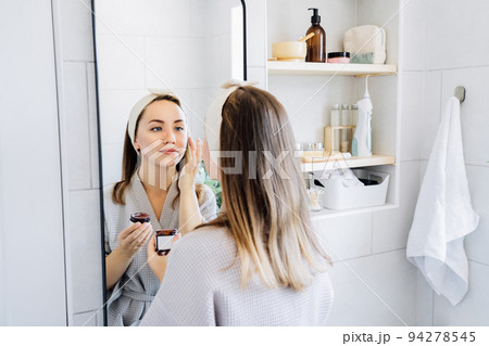 Young woman in bathrobe looking in the mirror and applying facial natural cosmetic clay mask on her face in bathroom. Cosmetic procedures for skin care at home. Beauty self-care. Selective focus. Young woman in bathrobe looking in the mirror and applying facial natural cosmetic clay mask on her face in bathroom. Cosmetic procedures for skin care at home. Beauty self-care. Selective focus. 94278545