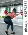 A girl with a sporty figure and a raised back performs bends with a barbell in front of a mirror in a fitness club. 94279882