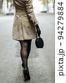 A girl in dark stockings and black stiletto shoes. She's wearing a beige trench coat, and she's carrying a small purse. Day walk in the city. 94279884