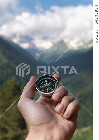 Magnetic compass in the palm of a male hand against the backdrop of a mountain range in the clouds in the summer outdoors, travel, first-person view Magnetic compass in the palm of a male hand against the backdrop of a mountain range in the clouds in the summer outdoors, travel, first-person view 94282924