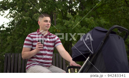 A young single father drinks coffee and rocks a stroller with a sleeping baby. A man with a stroller on a bench in the park. 94283341