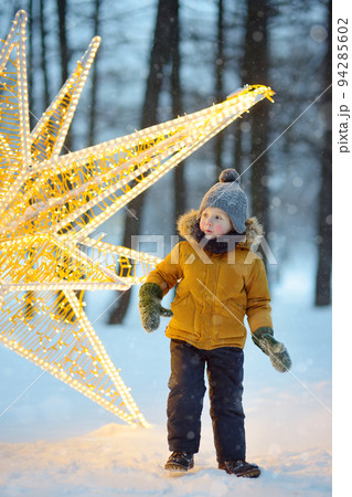 Little boy is admiring a large glowing street decoration in the form of a Christmas star. Traditional city Xmas market in the open air. Modern urban festive decoration. 94285602