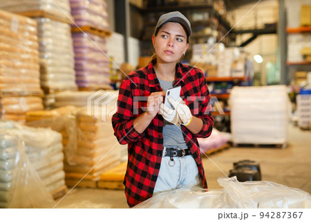 Female manager keeps records of bulk construction materials in the warehouse, writing down data on smartphone Female manager keeps records of bulk construction materials in the warehouse, writing down data on smartphone 94287367