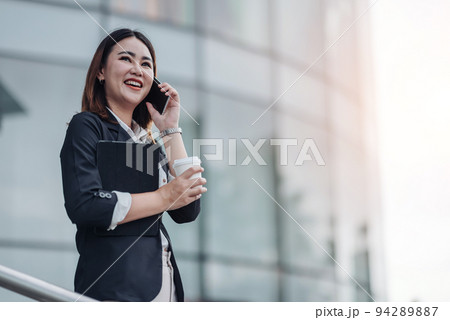 Young Asian businesswoman talking on phone and sitting in airport before business trip. Beautiful woman passenger has mobile call and discusses something with smile, holds coffee in hand 94289887