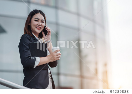 Young Asian businesswoman talking on phone and sitting in airport before business trip. Beautiful woman passenger has mobile call and discusses something with smile, holds coffee in hand Young Asian businesswoman talking on phone and sitting in airport before business trip. Beautiful woman passenger has mobile call and discusses something with smile, holds coffee in hand 94289888