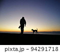 日暮れに海沿いを犬と散歩する人 94291718