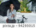 Young Asian businesswoman holding umbrella looking the watch and standing on a city street with bicycle looking at phone. 94293015