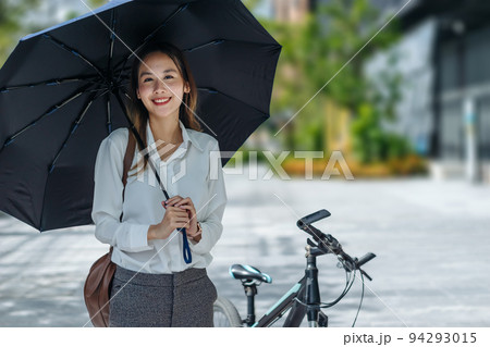 Young Asian businesswoman holding umbrella looking the watch and standing on a city street with bicycle looking at phone. Young Asian businesswoman holding umbrella looking the watch and standing on a city street with bicycle looking at phone. 94293015