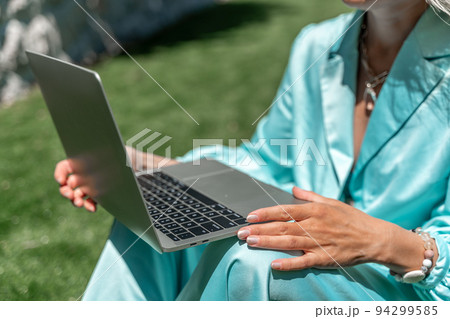 a young beautiful woman with blond curly hair in glasses and a blue dress sits on the grass in nature and uses a laptop, 94299585
