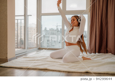 Side view portrait of relaxed woman listening to music with headphones lying on carpet at home. She is dressed in a white tracksuit. Side view portrait of relaxed woman listening to music with headphones lying on carpet at home. She is dressed in a white tracksuit. 94299644