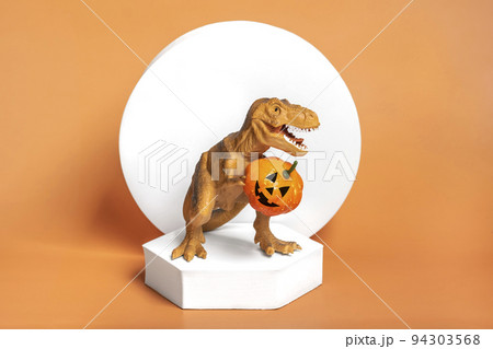Toy dinosaur Tyrannosaurus holding pumpkin in its paws, on white podium isolated on orange background Holiday greeting card Happy Halloween day, Hello Autumn creative minimal concept. Toy dinosaur Tyrannosaurus holding pumpkin in its paws, on white podium isolated on orange background Holiday greeting card Happy Halloween day, Hello Autumn creative minimal concept. 94303568