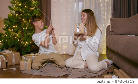 The younger brother takes pictures on his phone of his older sister with gifts at the Christmas tree The younger brother takes pictures on his phone of his older sister with gifts at the Christmas tree 94304218