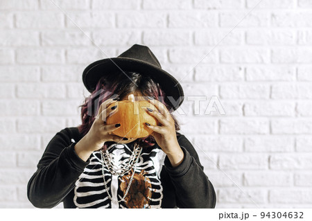 Young woman in costume witch with pumpkin head against white brick wall. teen dressed up as witch for celebrate Halloween. Young woman in costume witch with pumpkin head against white brick wall. teen dressed up as witch for celebrate Halloween. 94304632