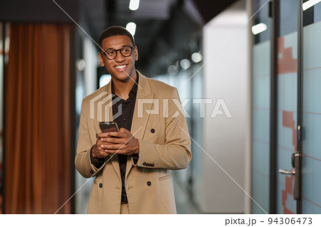Handsome smiling african businessman using phone while standing in modern coworking Handsome smiling african businessman using phone while standing in modern coworking 94306473