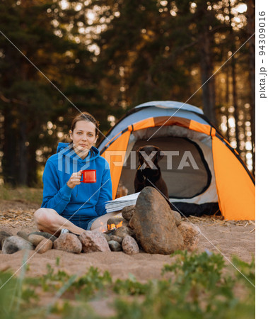 woman and a Labrador retriever dog on a camping trip with a tent in nature. a woman is reading a book or drinking tea from a red cup next to a campfire 94309016