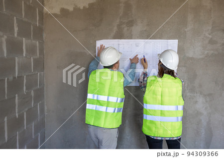 two engineers, a man and a woman in white helmets and protective vests, are standing in the room and holding a plan of the construction two engineers, a man and a woman in white helmets and protective vests, are standing in the room and holding a plan of the construction 94309366