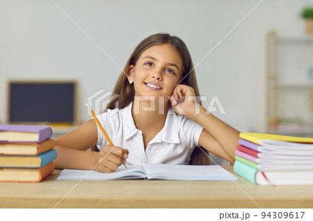 Portrait of funny and cute schoolgirl with dreamy expression sitting at desk between books. 94309617