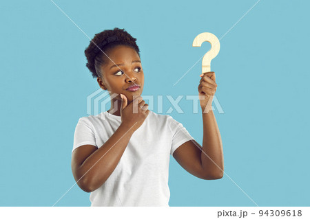 Young dark skinned woman with doubtful expression is looking at wooden question mark in her hand. 94309618