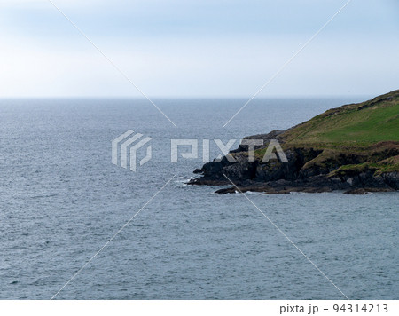 View of the coastal cliffs from the Warren beach. The rocky coast of the south of Ireland. Hills under the sky on a spring day, mountain beside sea under white clouds and sky. View of the coastal cliffs from the Warren beach. The rocky coast of the south of Ireland. Hills under the sky on a spring day, mountain beside sea under white clouds and sky. 94314213
