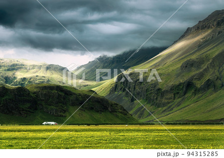 Dramatic Icelandic mountain with sunlight shining through storm clouds and house on field in summer at Iceland 94315285