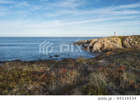 View of the sea and cliffs with the red and white lighthouse at praia pequena beach at wild Vicentina coast in Porto Covo, Portugal. Golden hour. 94316534