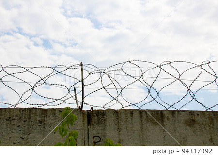 Barbed wire on the fence. Fencing stainless steel Barb Wire with sharp edges on a massive concrete wall for security on the background a blue sky. Barbwire on the prison fence. Military barbwire fence Barbed wire on the fence. Fencing stainless steel Barb Wire with sharp edges on a massive concrete wall for security on the background a blue sky. Barbwire on the prison fence. Military barbwire fence 94317092
