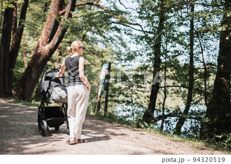 Rear view of casualy dressed mother walking with baby stroller in city park. Family, child and parenthood concept. 94320519