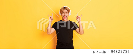 Joyful handsome asian guy enjoying listening music in headphones, showing rock-n-roll gesture, standing over yellow background Joyful handsome asian guy enjoying listening music in headphones, showing rock-n-roll gesture, standing over yellow background 94325752