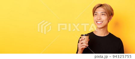 Close-up of relaxed and happy stylish asian blond guy, smiling and looking left dreamy while drinking coffee from takeaway cup, standing over yellow background 94325755