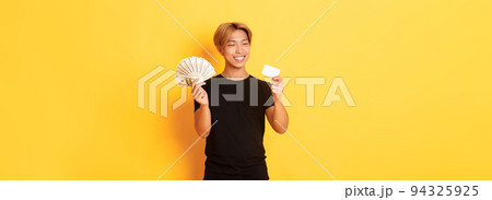 Portrait of satisfied smiling, handsome asian guy looking pleased at credit card while also holding money, standing yellow background 94325925