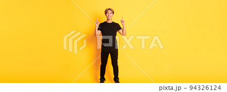 Full-length of confident handsome asian man with blond hair, smiling and pointing fingers up, showing logo, yellow background 94326124
