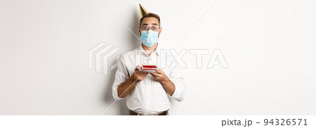 Covid-19, social distancing and celebration. Surprised birthday guy holding bday cake, wearing face mask from coronavirus, white background Covid-19, social distancing and celebration. Surprised birthday guy holding bday cake, wearing face mask from coronavirus, white background 94326571