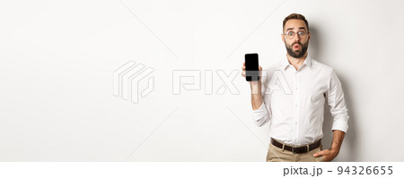 Surprised handsome manager in glasses, looking curious and showing mobile screen, standing over white background Surprised handsome manager in glasses, looking curious and showing mobile screen, standing over white background 94326655