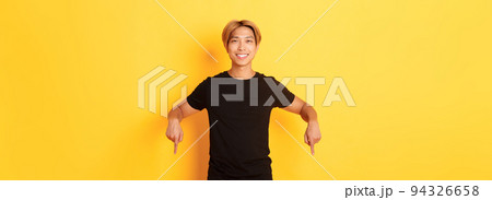 Portrait of smiling attractive korean guy with blond hair, wearing black outfit, pointing fingers down, showing way, standing yellow background Portrait of smiling attractive korean guy with blond hair, wearing black outfit, pointing fingers down, showing way, standing yellow background 94326658