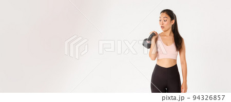 Sport, wellbeing and active lifestyle concept. Portrait of cute brunette asian fitness girl, sign up bodybuilding classes at gym, surprised with weight of kettlebell, standing over white background 94326857