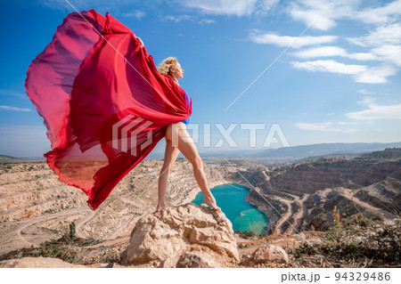 Side view of a beautiful sensual woman in a red long dress posing on a rock high above the lake in the afternoon. Against the background of the blue sky and the lake in the form of a heart Side view of a beautiful sensual woman in a red long dress posing on a rock high above the lake in the afternoon. Against the background of the blue sky and the lake in the form of a heart 94329486