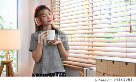 Calm young woman holding cup of coffee and listening yo music on wireless headphone, enjoy stress free peaceful mood 94331063