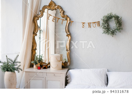 The interior of a bedroom decorated for Christmas and New Year in a Scandinavian or vintage style. A mirror over the dresser, spruce branches in a vase, a Christmas wreath over the bed 94331678