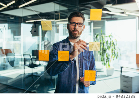 Focused senior bearded businessman in casual clothes writing on sticker with black marker. Colorful notes stuck on a glass wall in a conference room. Business, planning and management concept Focused senior bearded businessman in casual clothes writing on sticker with black marker. Colorful notes stuck on a glass wall in a conference room. Business, planning and management concept 94332338