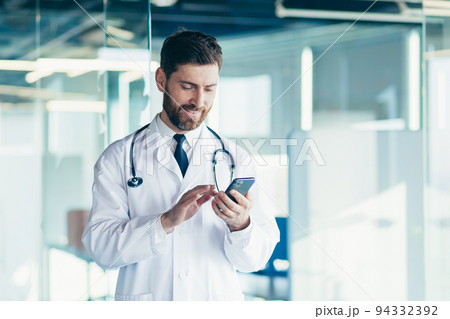 Male doctor in a white coat in a modern clinic reads information from a mobile phone corresponds with colleagues happy and smiles 94332392