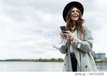 portrait of a smiling young woman in a black hat and autumn gray coat with a phone on the background 94332805