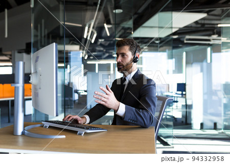 Business man in a modern office works at a computer, uses a headset for video calling, technical support Business man in a modern office works at a computer, uses a headset for video calling, technical support 94332958