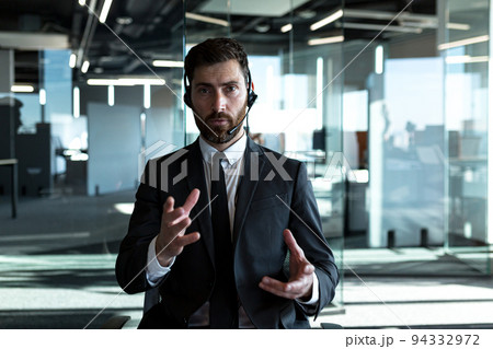 The employee makes a video call, looks at the camera and explains the task, serious gestures with his hands works in a modern office, uses a headset The employee makes a video call, looks at the camera and explains the task, serious gestures with his hands works in a modern office, uses a headset 94332972