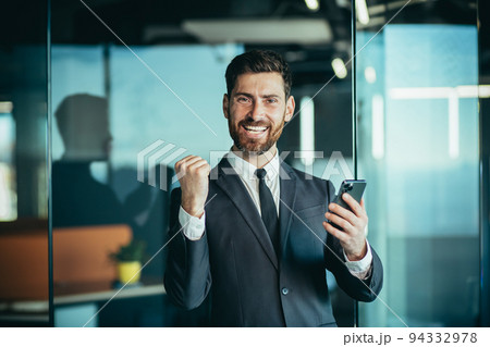 Happy businessman boss looks at camera and smiles, holds phone in hands, man wins online and celebrates victory Happy businessman boss looks at camera and smiles, holds phone in hands, man wins online and celebrates victory 94332978