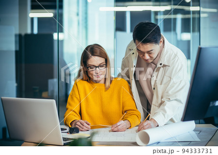 Two employees of designers and architects, thinking about the project, an Asian man and a blonde woman, in casual clothes working together at the table, with a plan drawing project 94333731