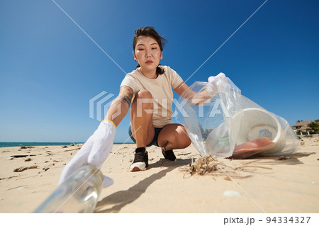 Serious female volunteer collecting garbage on sandy beach Serious female volunteer collecting garbage on sandy beach 94334327
