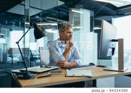 A gray-haired man thinks about the tasks, a businessman with a beard works in a modern office at the computer, 94334390