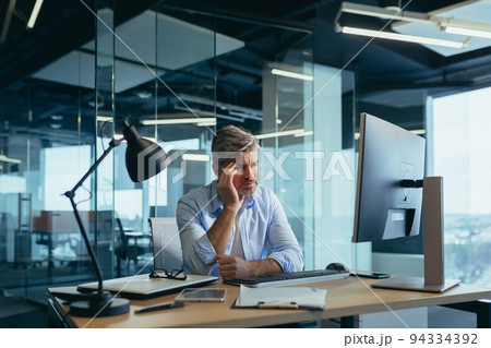 An experienced gray-haired businessman works at the computer until late, tired has a severe headache, stress and depression 94334392