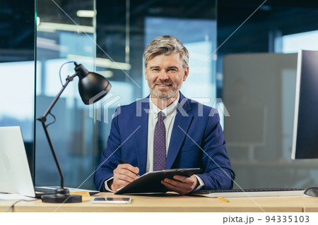 business owner looks at the camera and smiles, gray-haired man signs documents, businessman on paper works at the computer in a modern office business owner looks at the camera and smiles, gray-haired man signs documents, businessman on paper works at the computer in a modern office 94335103