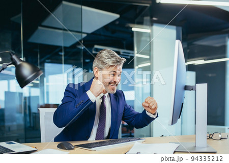 Successful boss works in a modern office at the computer, a senior man gray and happy with a beard looks at the camera and smiles, holds his hands up, celebrates victory Successful boss works in a modern office at the computer, a senior man gray and happy with a beard looks at the camera and smiles, holds his hands up, celebrates victory 94335212